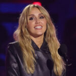 Miley Cyrus, cheveux blonds et veste en cuir noir, souriante au micro lors des iHeartRadio Music Awards 2026